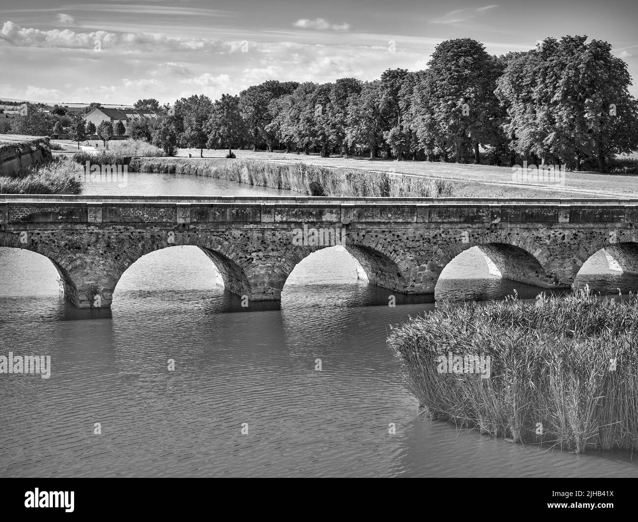 Schwarzweißansicht einer alten gewölbten Steinbrücke über das Wasser in Holic, Slowakei, mit ruhigem Fluss, Schilfrohr und historischer Landschaft Stockfoto