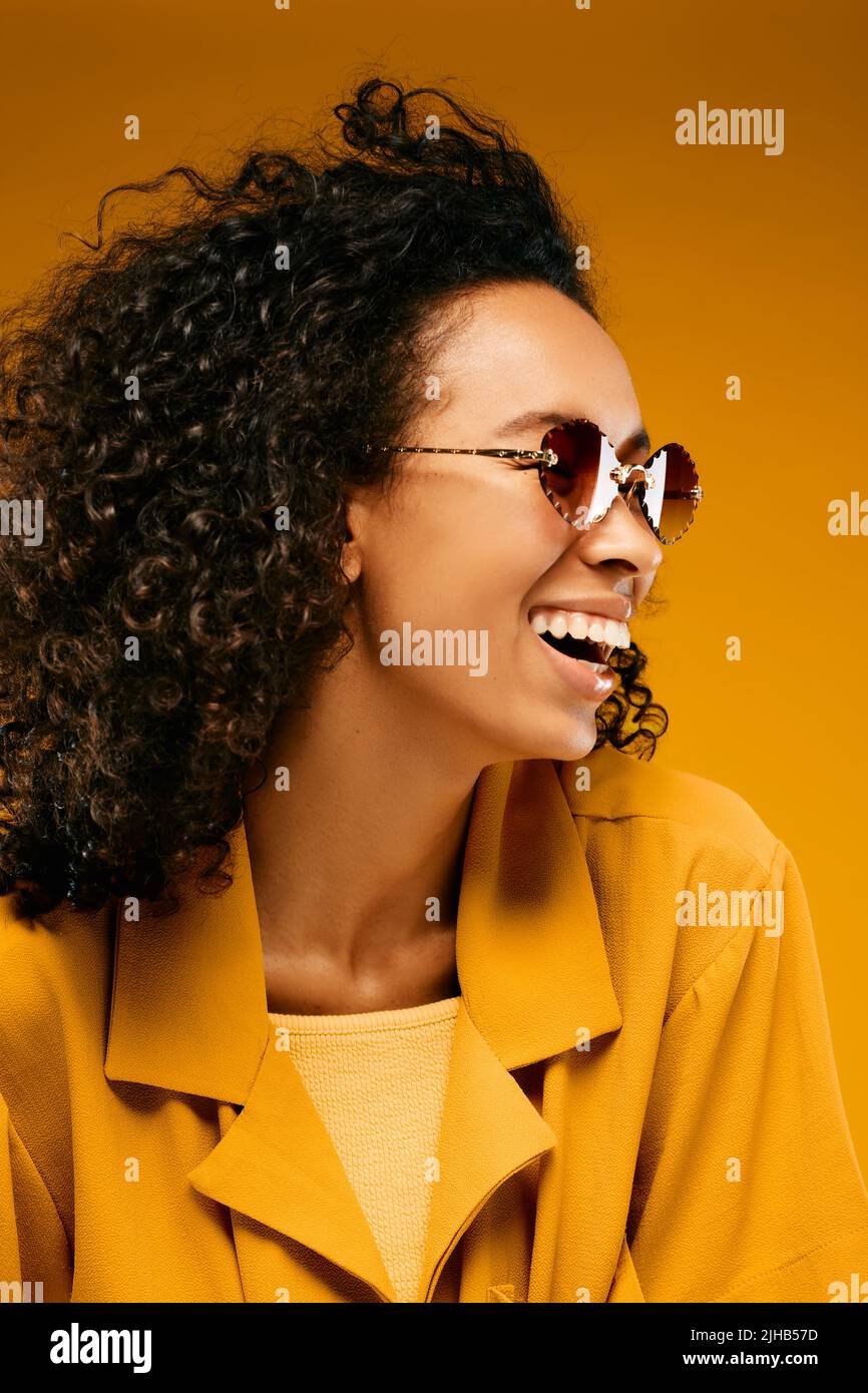 Lockiges afroamerikanisches Mädchen, das in Sonnenbrillen und gelber Kleidung auf gelbem Hintergrund getragen wird und fröhlich lacht Stockfoto