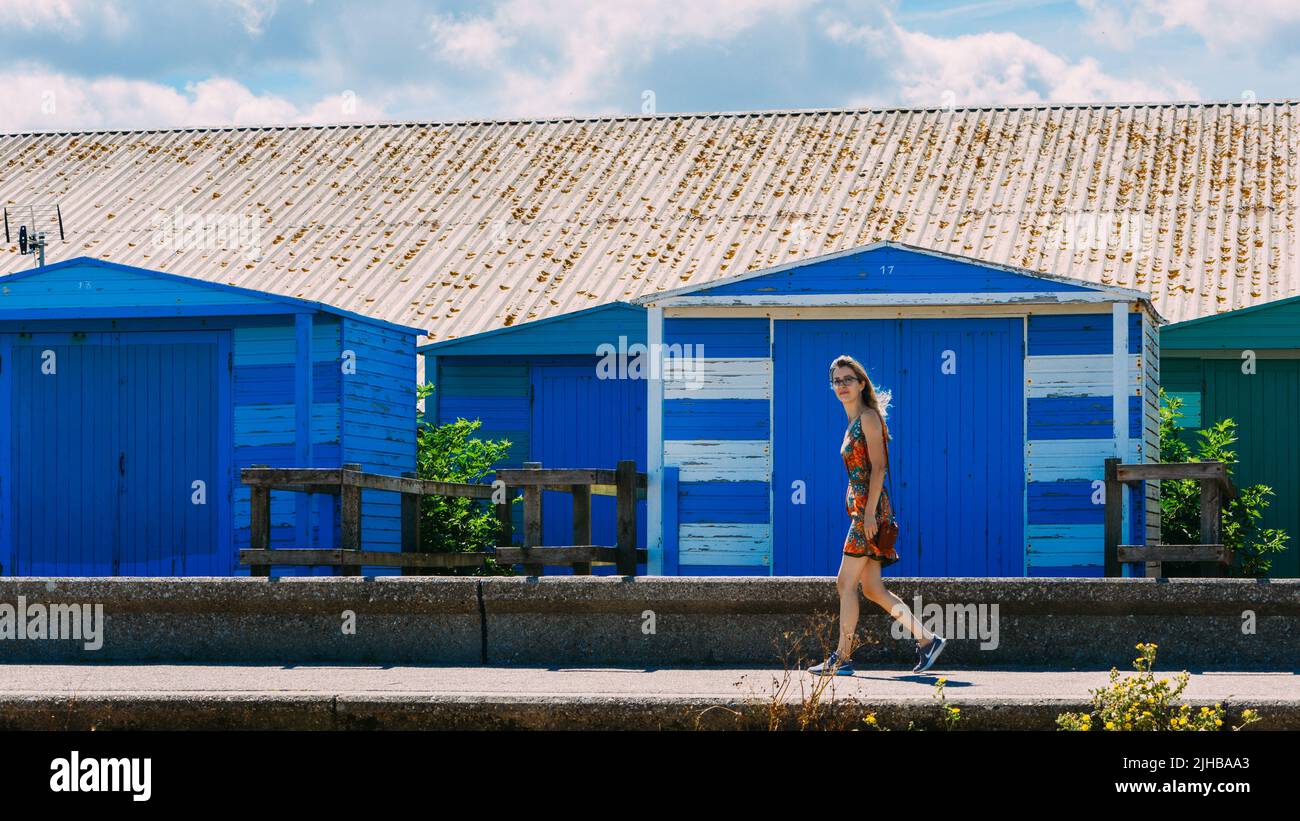 Junge Frau geht neben einer strahlend blauen Strandhütte in Whitstable, Kent, Großbritannien Stockfoto