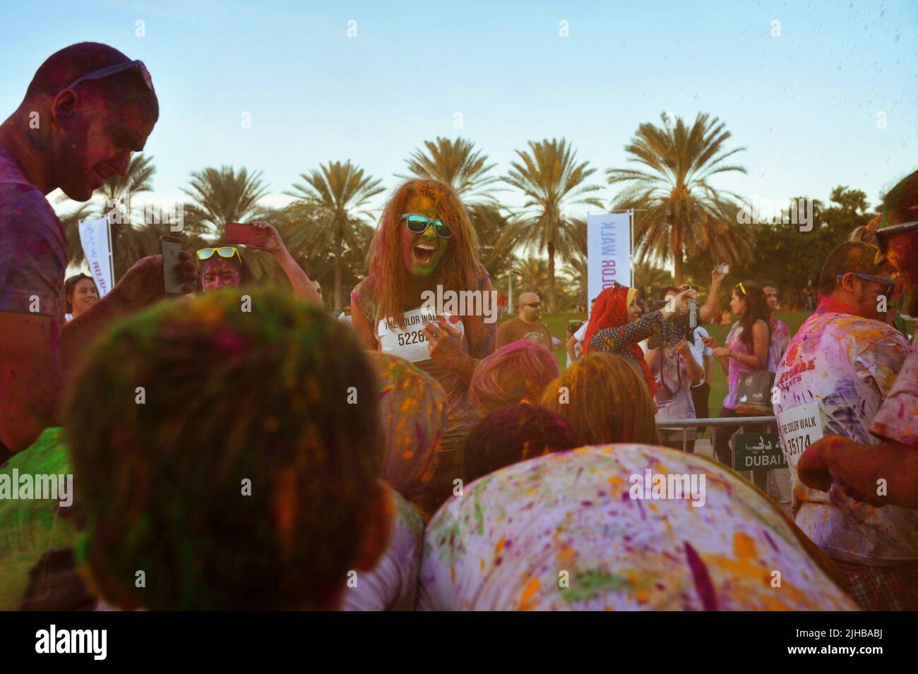 Die junge kaukasische Frau lacht herzlich, als sie das Chaos um sie herum sieht: Eine Menge schnappte sich beim Color Walk in Dubai, VAE, mehr buntes Pulver. Stockfoto