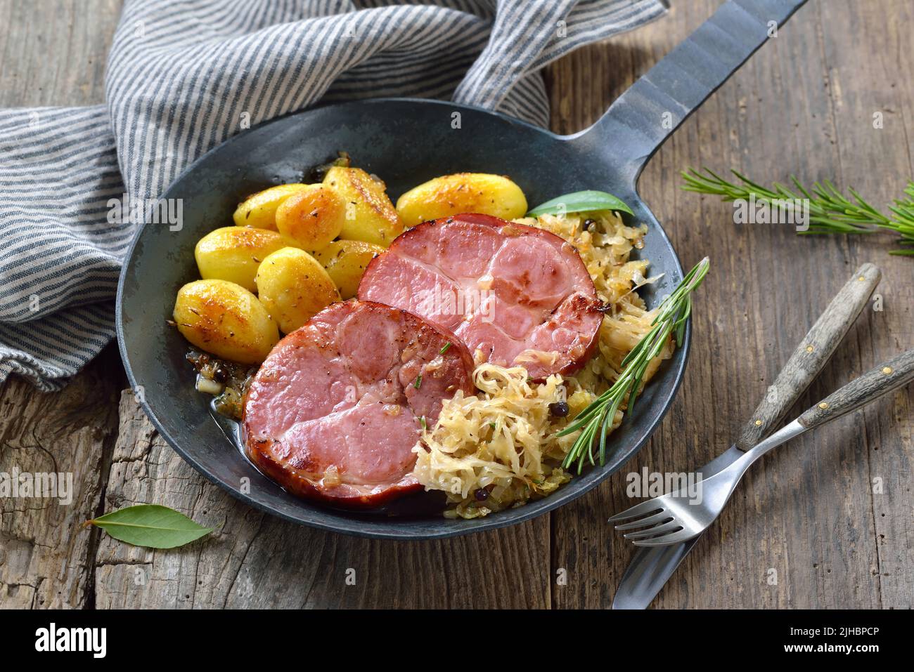 Deutsche Küche – geräucherter Schweinehalk mit Sauerkraut und gebratenen Babykartoffeln, die in einer eisernen Pfanne auf einem alten Holztisch serviert werden Stockfoto