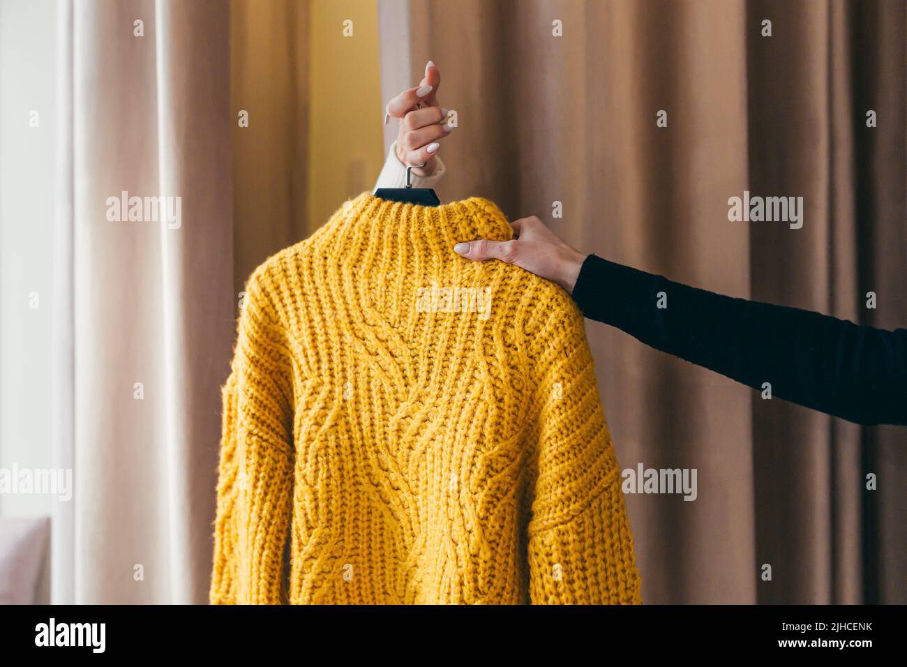 Nahaufnahme. Die Hand eines Verkäufers oder Beraters, der Kleidung, einen gelben Pullover auf einem Kleiderbügel, an die Hand eines Kunden übergibt, der in der Umkleidekammer hinter den Vorhängen wartet. In einem Bekleidungsgeschäft in einem Einkaufszentrum Stockfoto