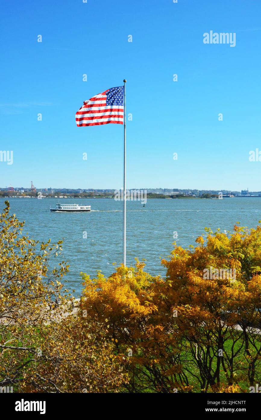 NEW YORK, NY - 04 NOV 2019: Fahnenmast mit Fallbäumen auf Ellis Island mit einem Statue Cruises Boot auf dem Fluss Stockfoto
