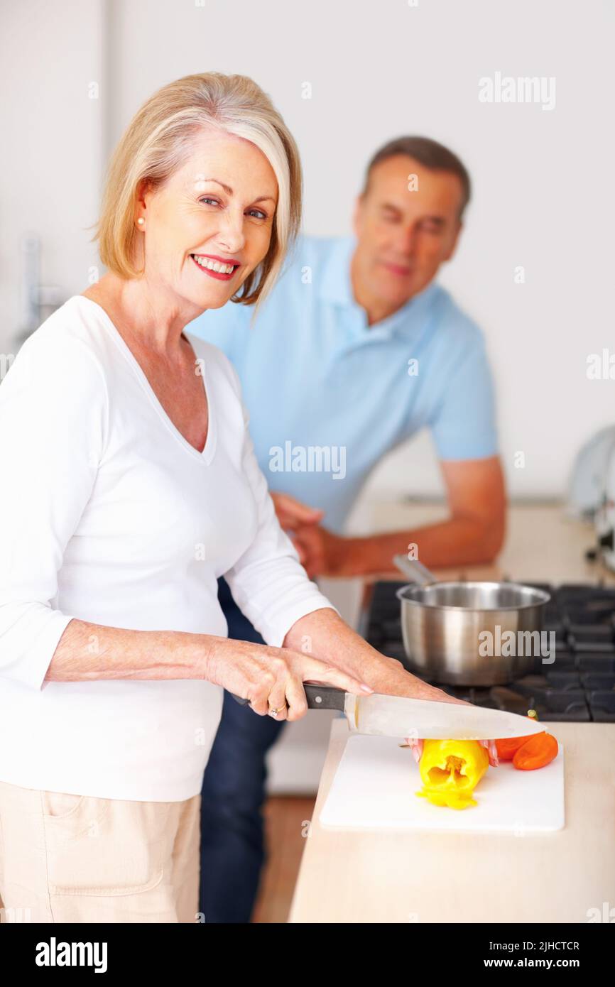 Lächelnde ältere Frau, die mit einem Mann im Hintergrund Essen zubereitete. Porträt einer lächelnden reifen Frau, die mit einem Mann im Hintergrund in der Küche Essen zubereitet. Stockfoto