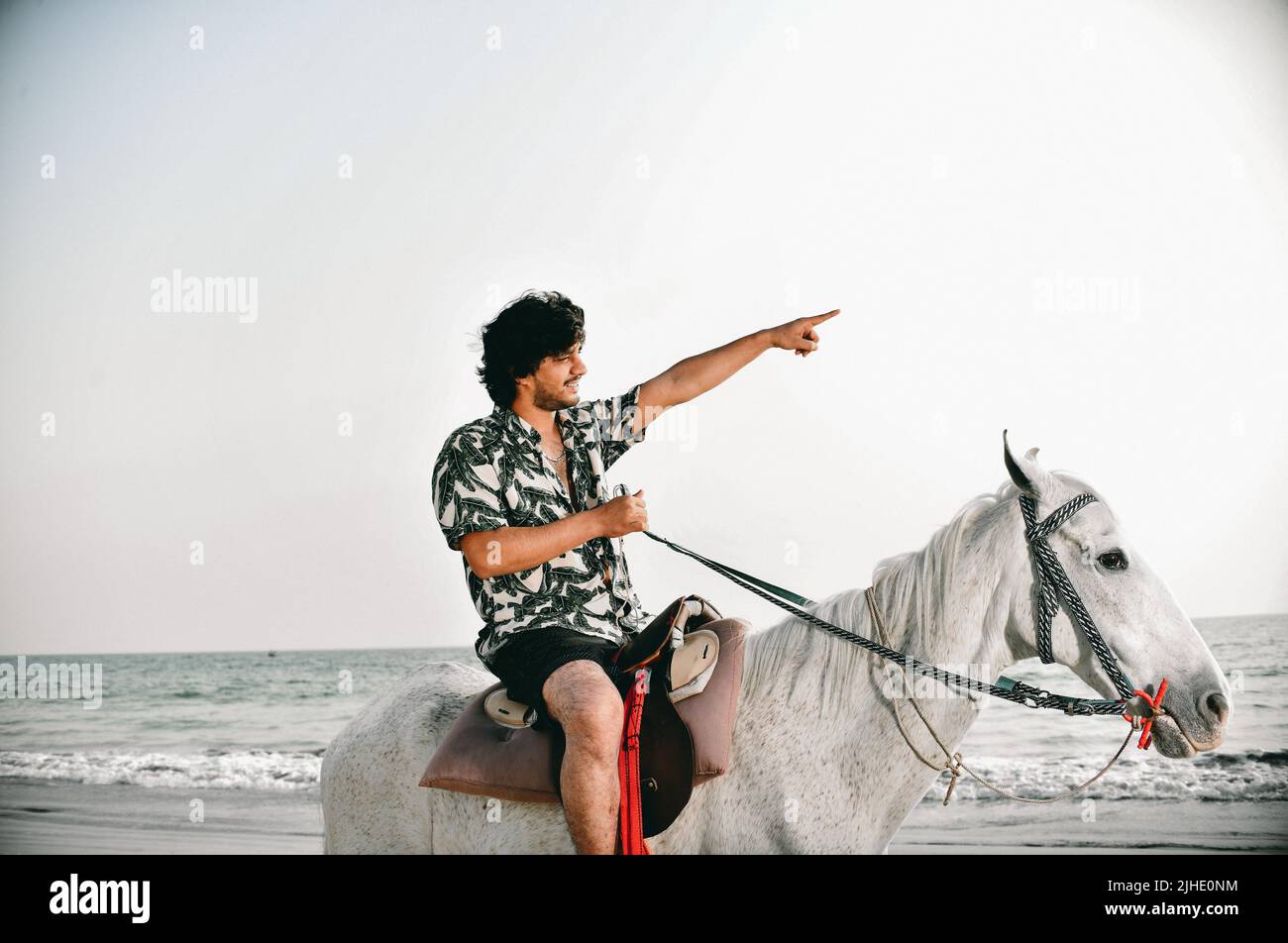 Hübscher Junge, der am Strand auf einem Pferd sitzt und mit der Hand die Richtung zeigt Stockfoto