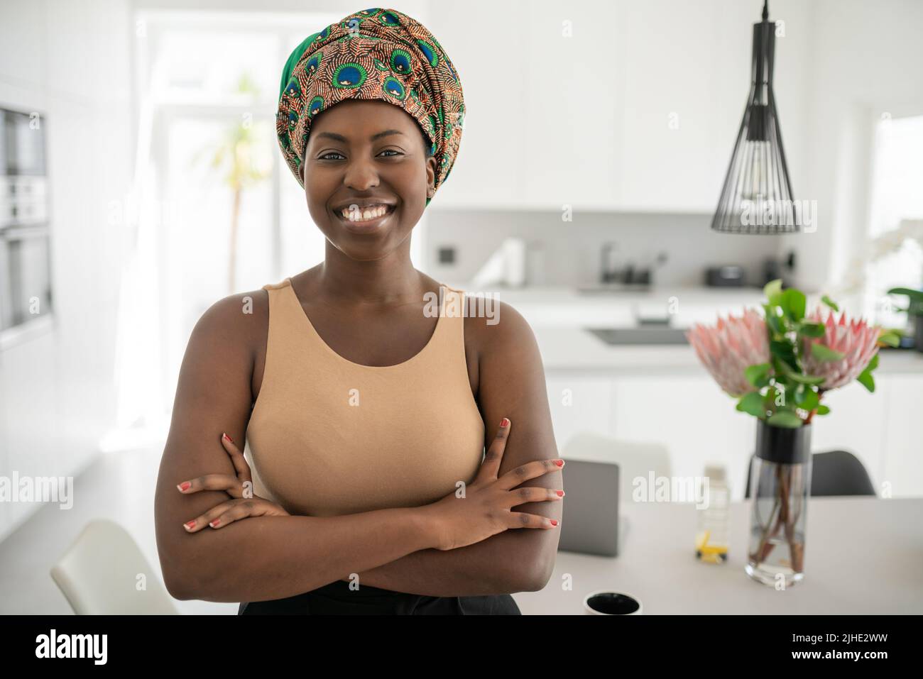 Porträt einer selbstbewussten, lächelnden Afrikanerin mit gekreuzten Armen und Blick in die Kamera. Arbeiten von zu Hause aus und tragen ein traditionelles Kopftuch Stockfoto