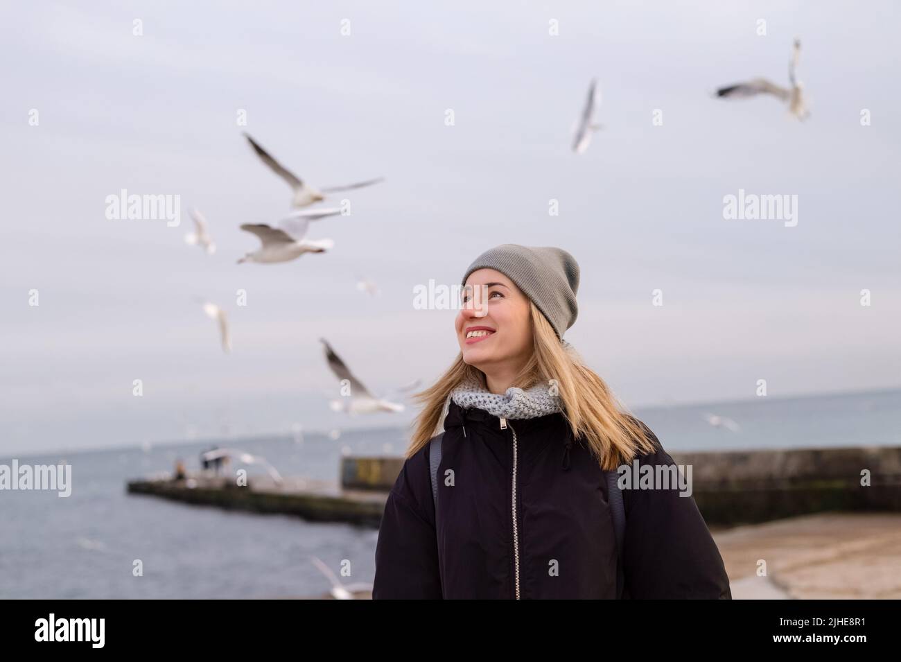 Lächelnde, hübsche junge Frau am Winterstrand, die Möwen ansieht Stockfoto