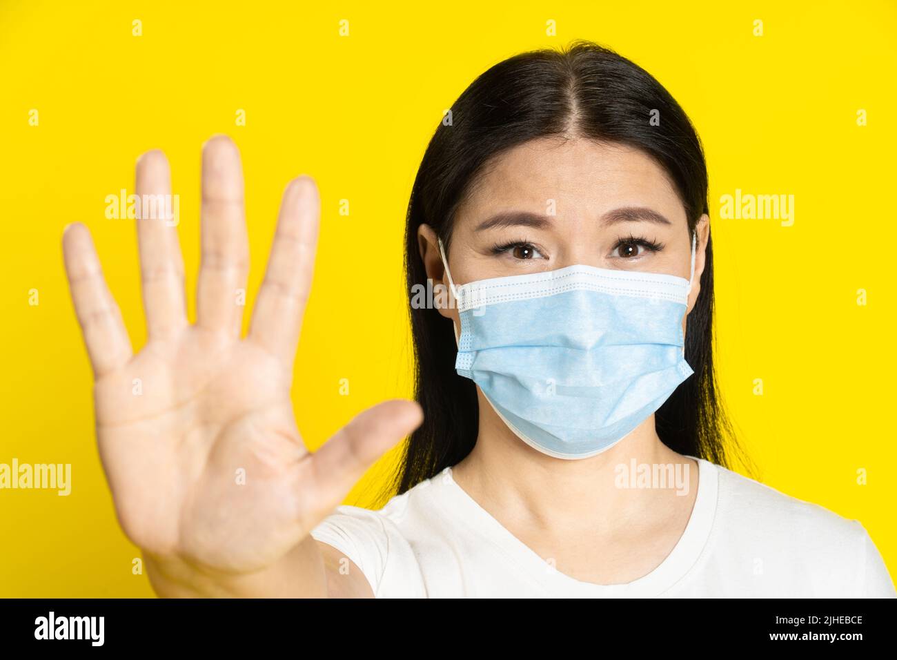 Bitten, mittleren Alters asiatische Frau tragen medizinische Maske Gestik zu stoppen, um mit positiven Augen auf Kamera isoliert auf gelbem Hintergrund zu halten. Pandemiekonzept, Affenpocken- oder Coronavirus. Stockfoto