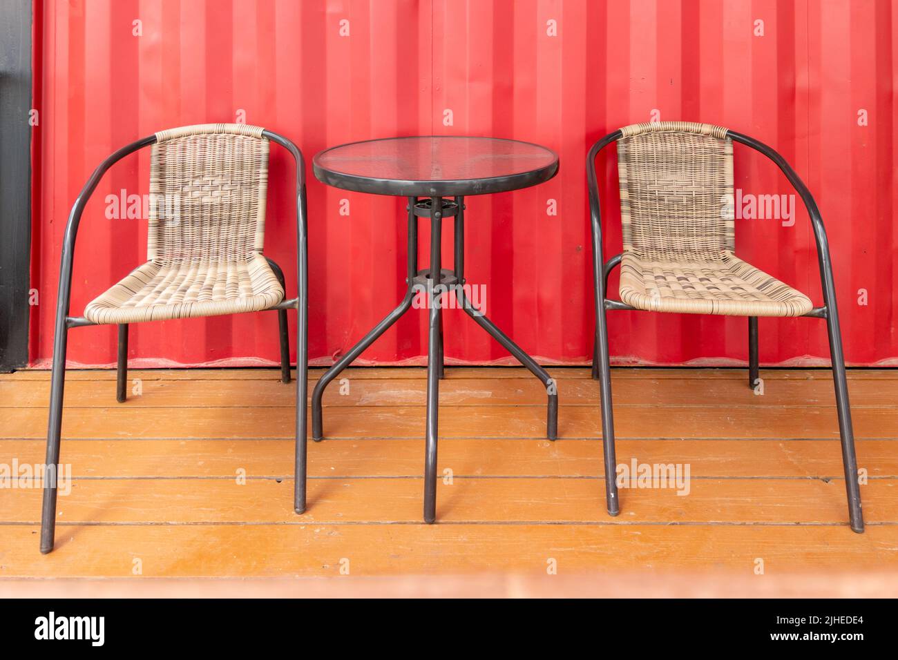 Zwei Korbsessel und Glastisch in der Nähe der roten Wand des Straßencafés, leer Stockfoto