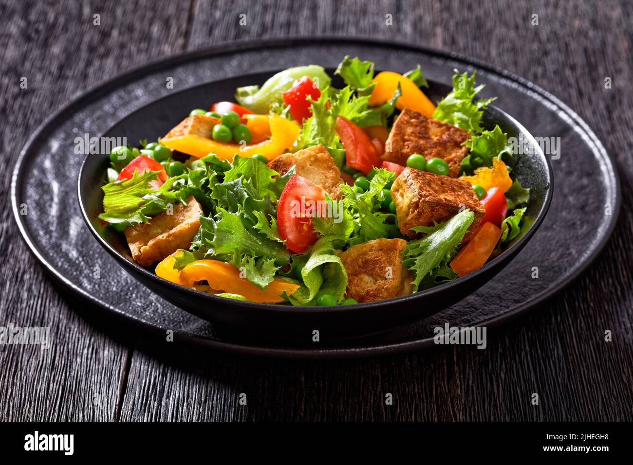 Gebratener Tofu-Salat mit knackigen Salatblättern, grünen Erbsen, Tomaten, gelbem Paprika in schwarzer Schüssel Stockfoto