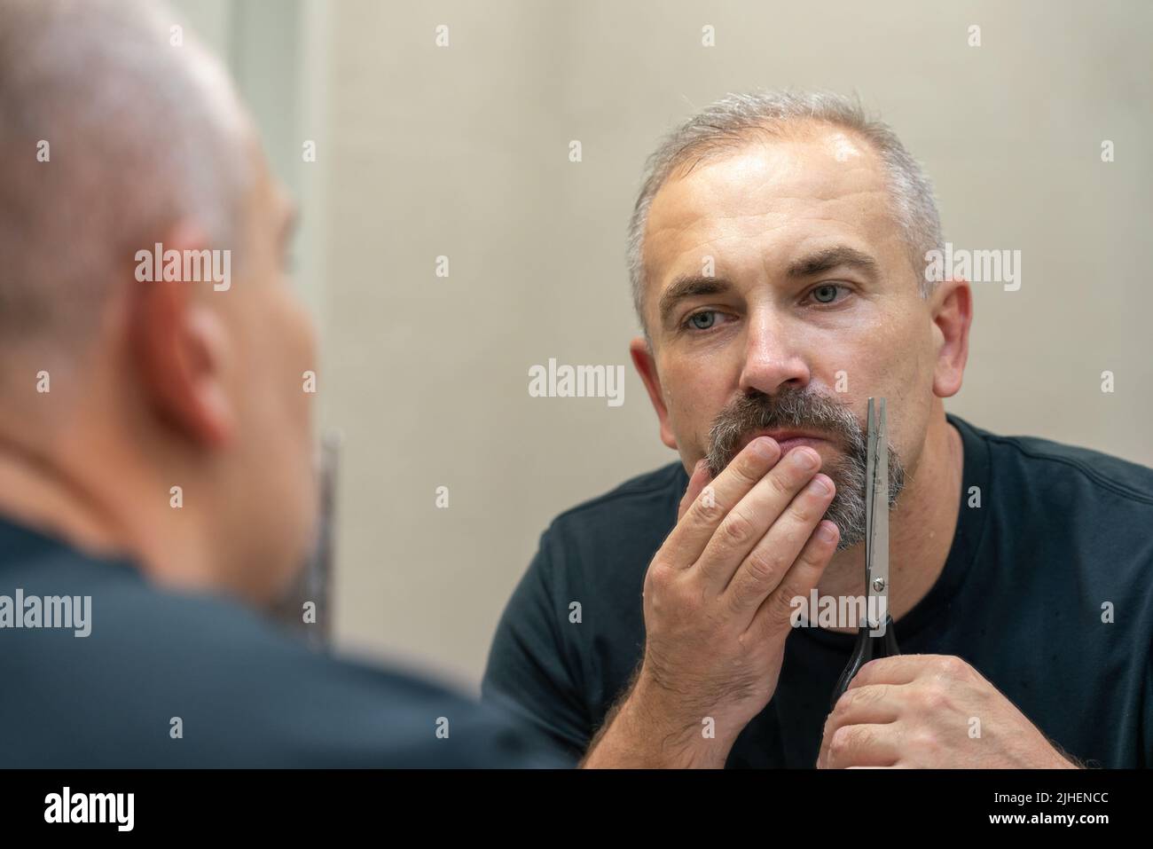 Ein gutaussehender Mann mittleren Alters, der mit einer Schere seinen Bart ein bisschen abschneidet und die Form fixiert Stockfoto