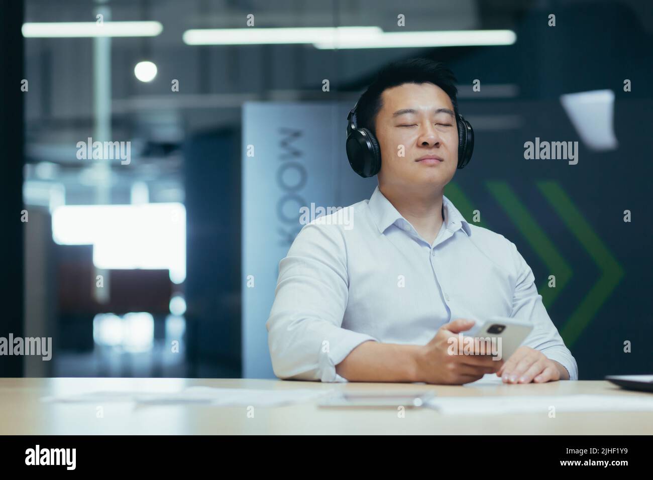Der asiatische Chef-Unternehmer entspannt sich im Büro, hört entspannende und entspannende Musik, Mann sitzt am Schreibtisch, mit großen Kopfhörern und Online-Höranwendungen am Telefon Stockfoto