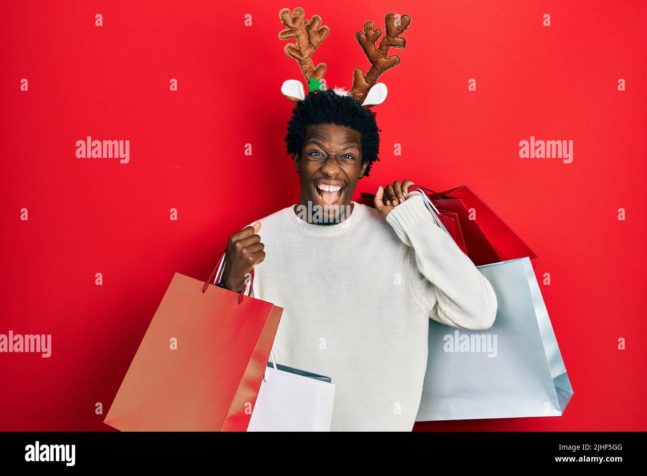 Junger afroamerikanischer Mann mit Hirsch-weihnachtshut und Einkaufstaschen, der verrückt und erstaunt über den Erfolg feierte und aufgeregt mit offenen Augen schrie. Stockfoto