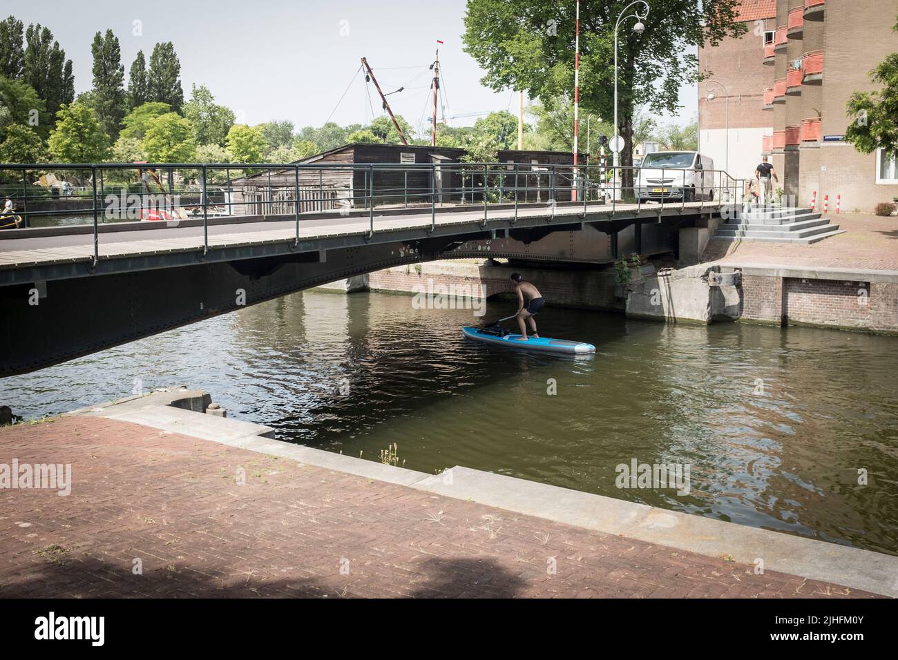 Ein Mann auf einem Stehpaddelbrett duckt an einem heißen und sonnigen Tag in Amsterdam, Niederlande, unter einer Brücke auf einem Kanal. Stockfoto