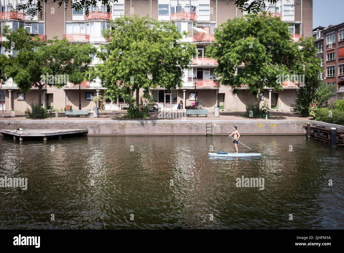 An einem heißen und sonnigen Tag in Amsterdam, Niederlande, paddelt ein Mann auf einem Stand-up-Paddle-Board an einem Apartmentgebäude auf einem Kanal vorbei. Stockfoto