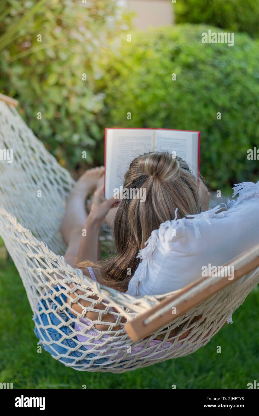 Vertikale Ansicht der jungen unkenntlichen Frau von hinten, die ein Buch liest, während sie im Sommer in einer Hängematte sitzt Stockfoto