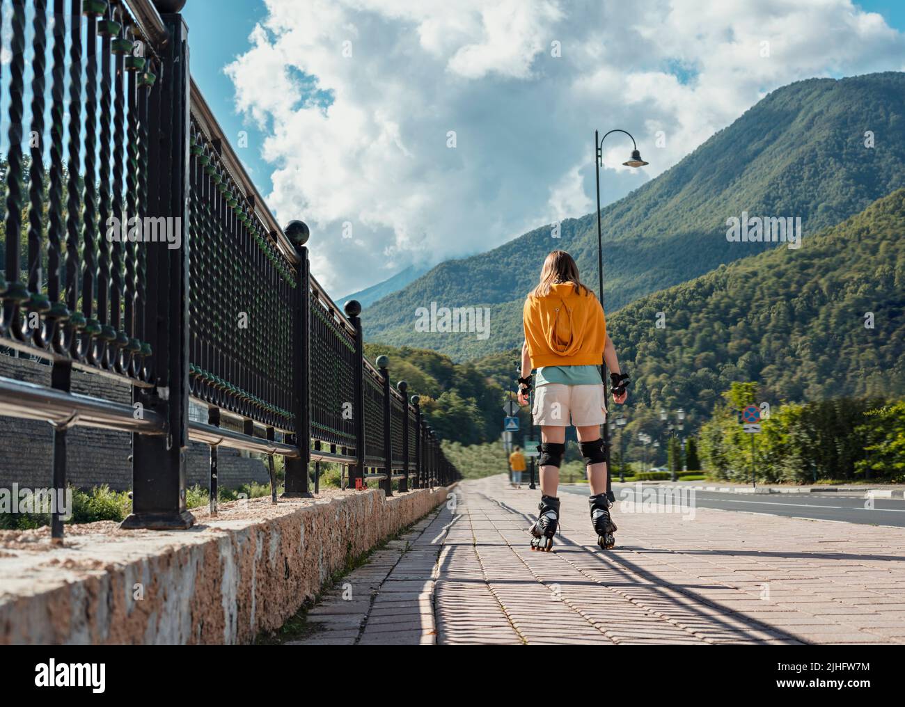 Rückansicht der jungen blonden Frau in Schutzausrüstung Reiten auf Rollschuhe entlang Böschung im Sommer gegen Berge aktiven Lebensstil, im Freien Stockfoto