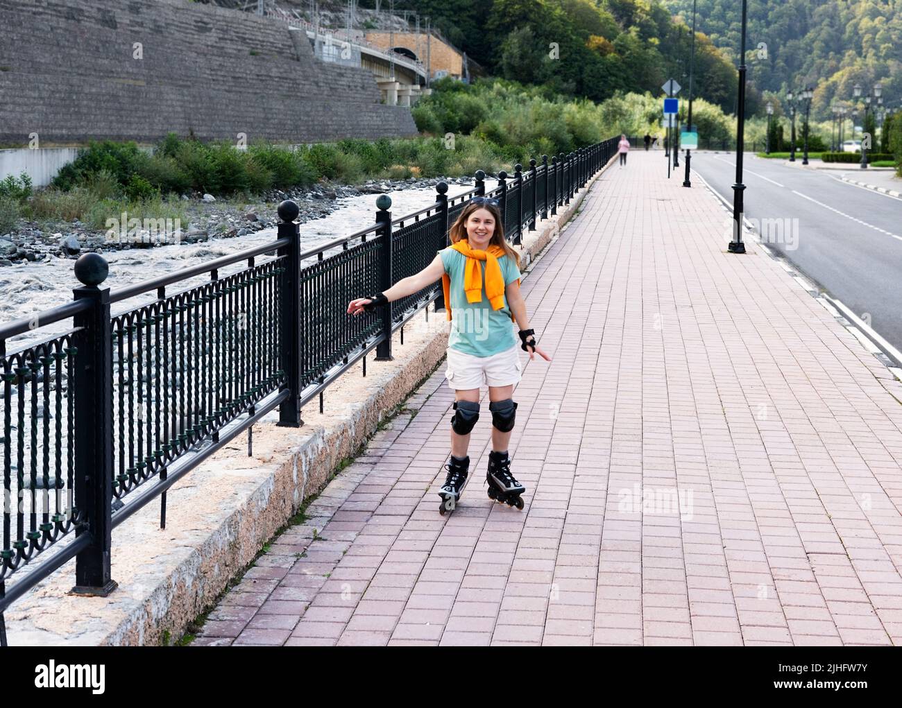 Glückliche schöne junge Frau in Schutzausrüstung Reiten auf Rollschuhe entlang Böschung im Sommer Outdoor-Aktivitäten, Rollschuhlaufen Stockfoto