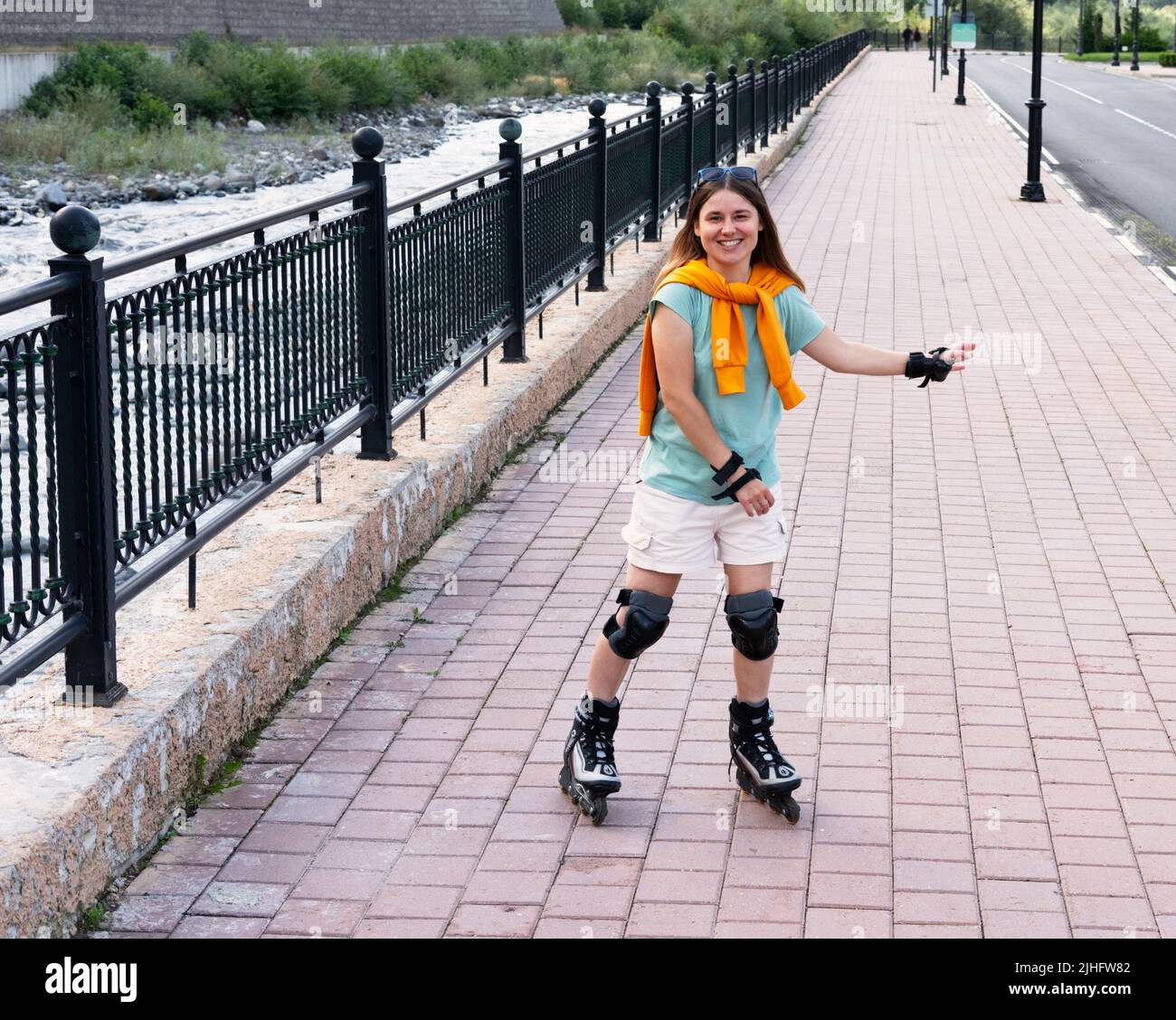 Glückliche schöne junge Frau in Schutzausrüstung Reiten auf Rollschuhe entlang Böschung im Sommer Outdoor-Aktivitäten, Rollschuhlaufen Stockfoto