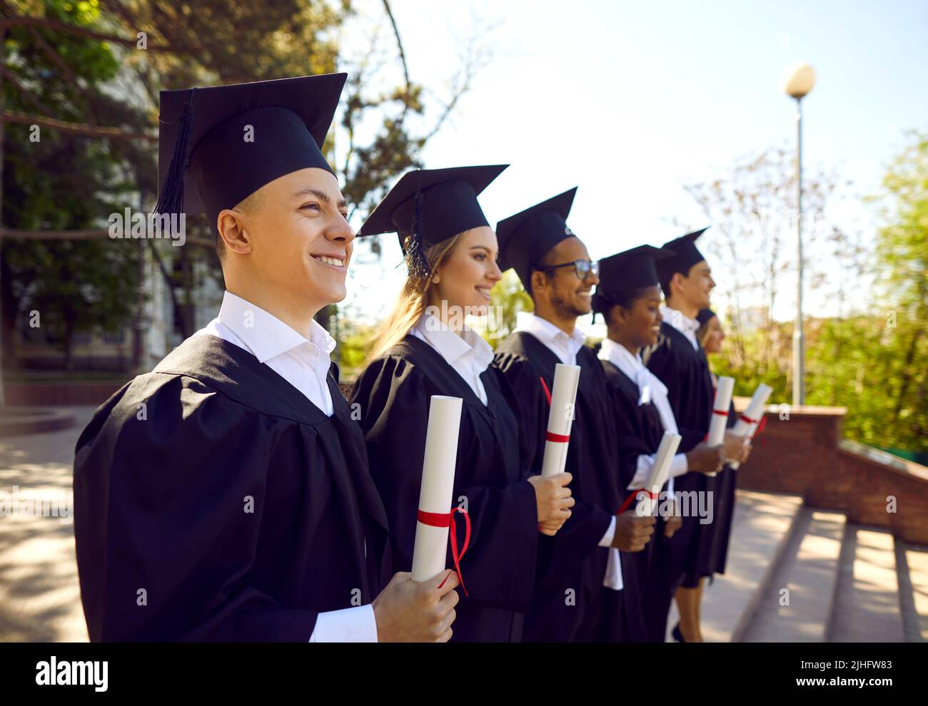 Glückliche multirassische Absolventen im Studentenkleid stehen während der Abschlussfeier der Universität in Reihe Stockfoto