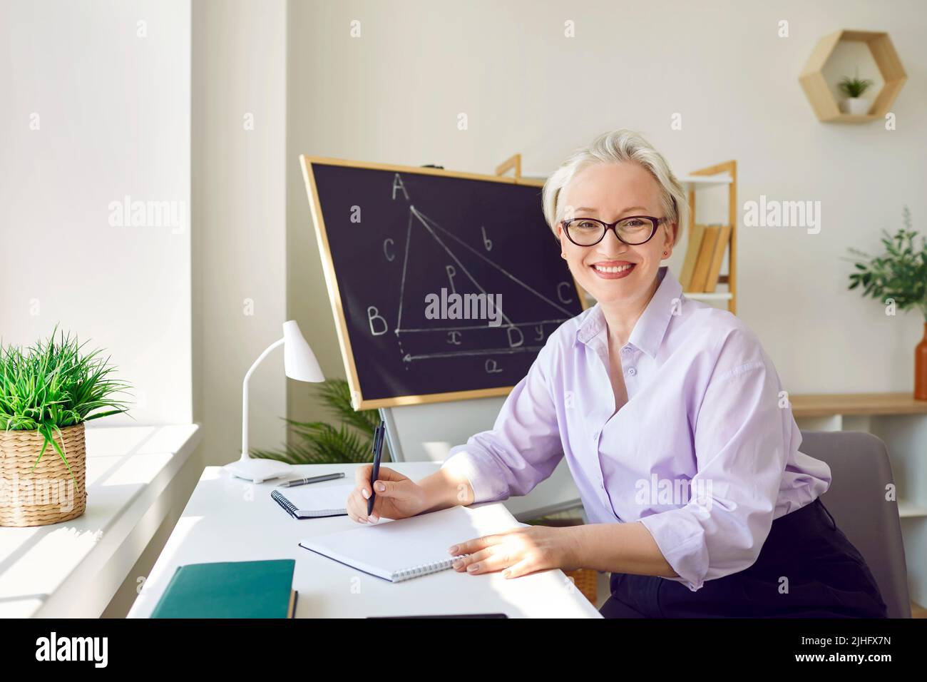 Glückliche Frau, die in der Schule als Mathematiklehrerin arbeitet und an ihrem Schreibtisch im Klassenzimmer sitzt Stockfoto