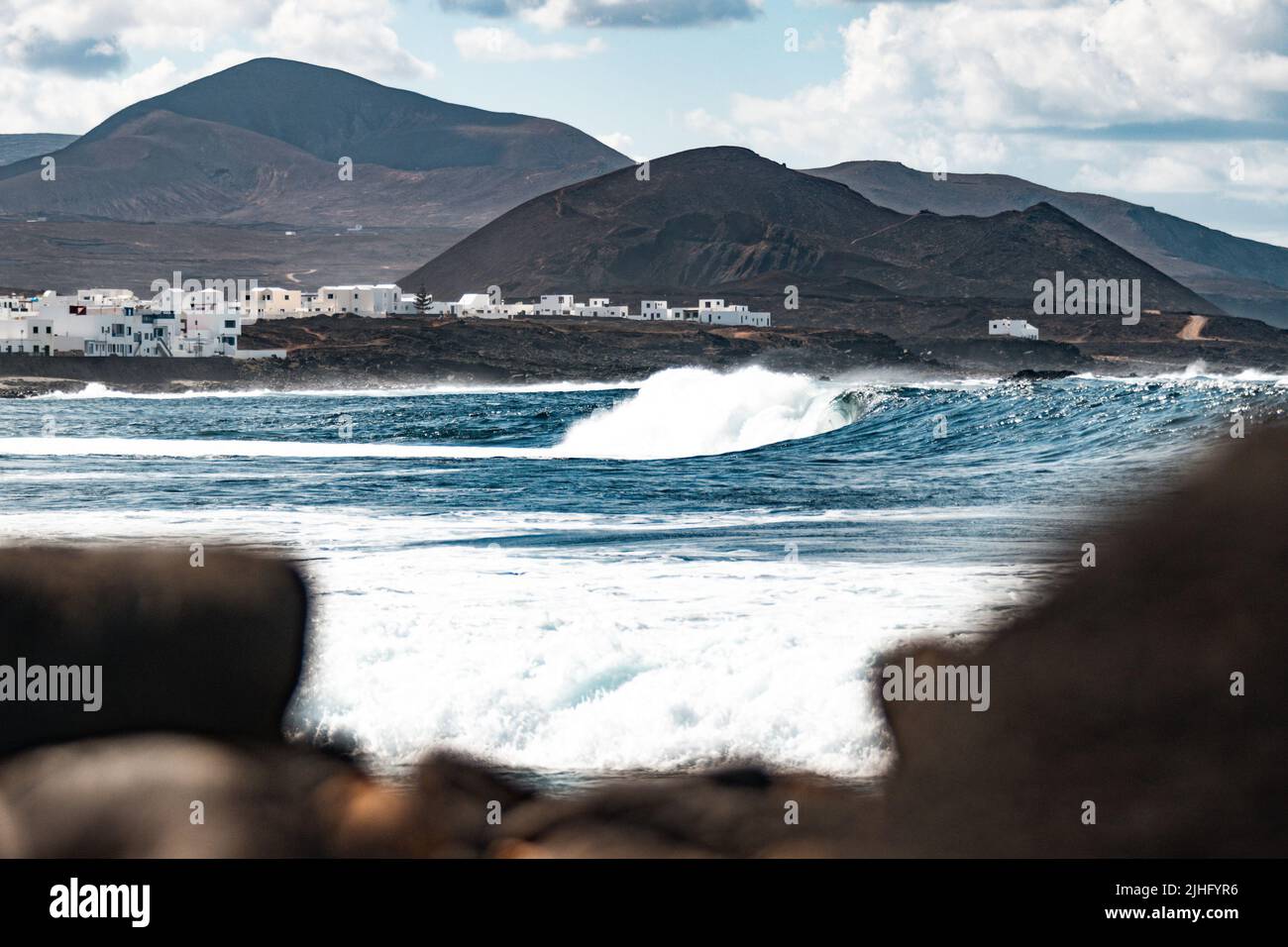 Wilde felsige Küste des Surfspots La Santa Lanzarote, Kanarische Inseln, Spanien. Surfer reiten eine große Welle in felsiger Bucht, Vulkan Berg im Hintergrund. Stockfoto