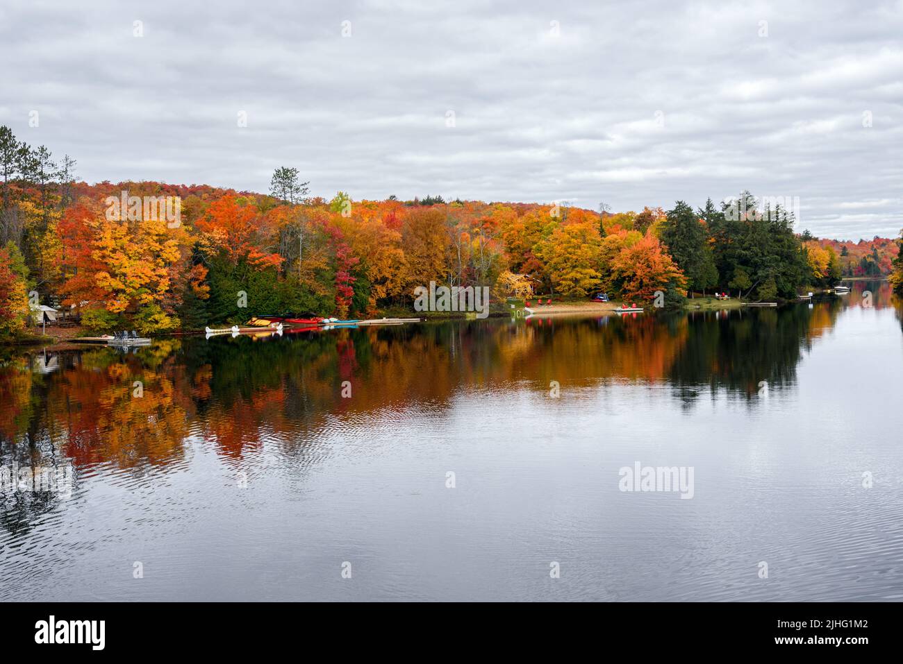 Hölzerne Stege mit Adirondack-Stuhl auf ihnen und bunte Kanus entlang der bewaldeten Ufer eines Sees an einem bewölkten Herbsttag, atemberaubende Herbstlaub. Stockfoto