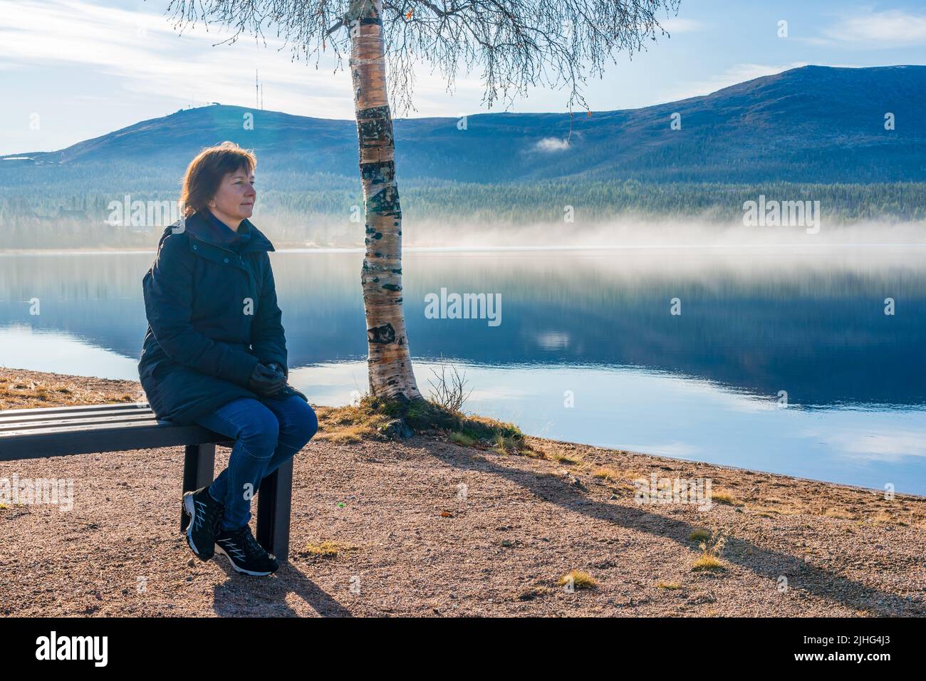 Frau, die auf einer Bank am Strand sitzt und die schöne Aussicht auf Berg und Fluss beobachtet, Gällivare, Schwedisch Lappland, Schweden Stockfoto