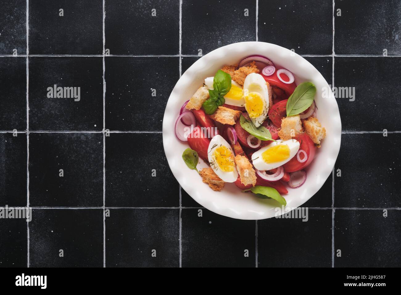 Toskanischer Panzanella-Salat. Frischer Tomaten-Salat mit Basilikumblättern, Eiern, Olivenöl und Zwiebeln in einer weißen Schüssel auf altem schwarzen Hintergrund. Vegetarisches Panzanell Stockfoto