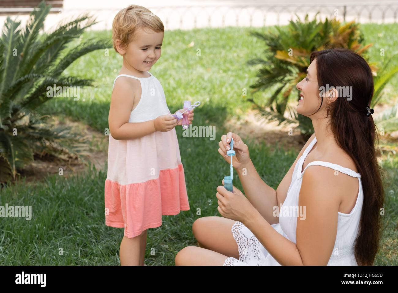 Lächelndes Kind hält Seifenblasen in der Nähe von Mama auf grünem Rasen Stockfoto