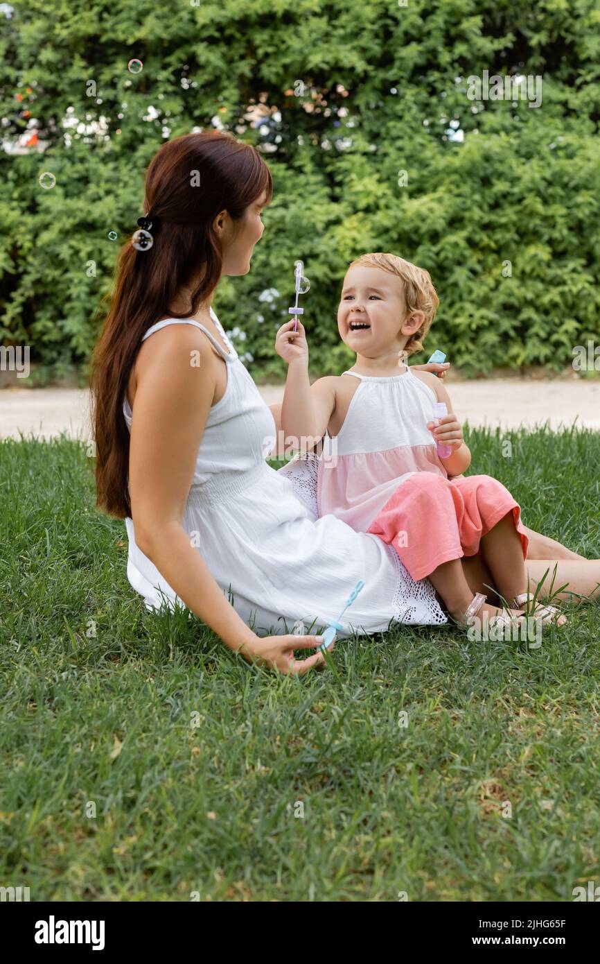 Fröhliches Baby Mädchen hält Seifenblasen in der Nähe Mama auf Glas im Park Stockfoto