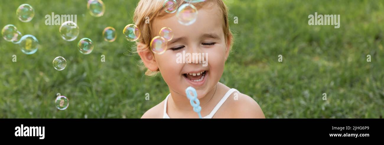Fröhliches Baby Mädchen hält Zauberstab in der Nähe Seifenblasen im Park, Banner Stockfoto
