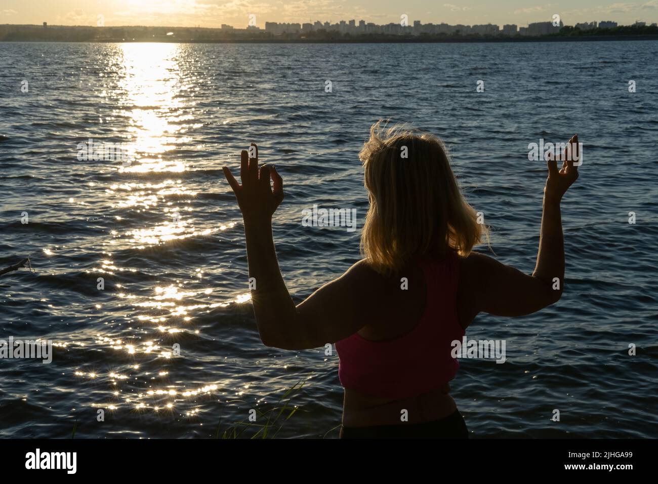 Silhouette einer Frau, die ihre Finger in einer Mudra-Pose vor dem Hintergrund des Wassers des Meeres, des Sees, hält. Meditation. Sport im Alter. Yoga ein Stockfoto
