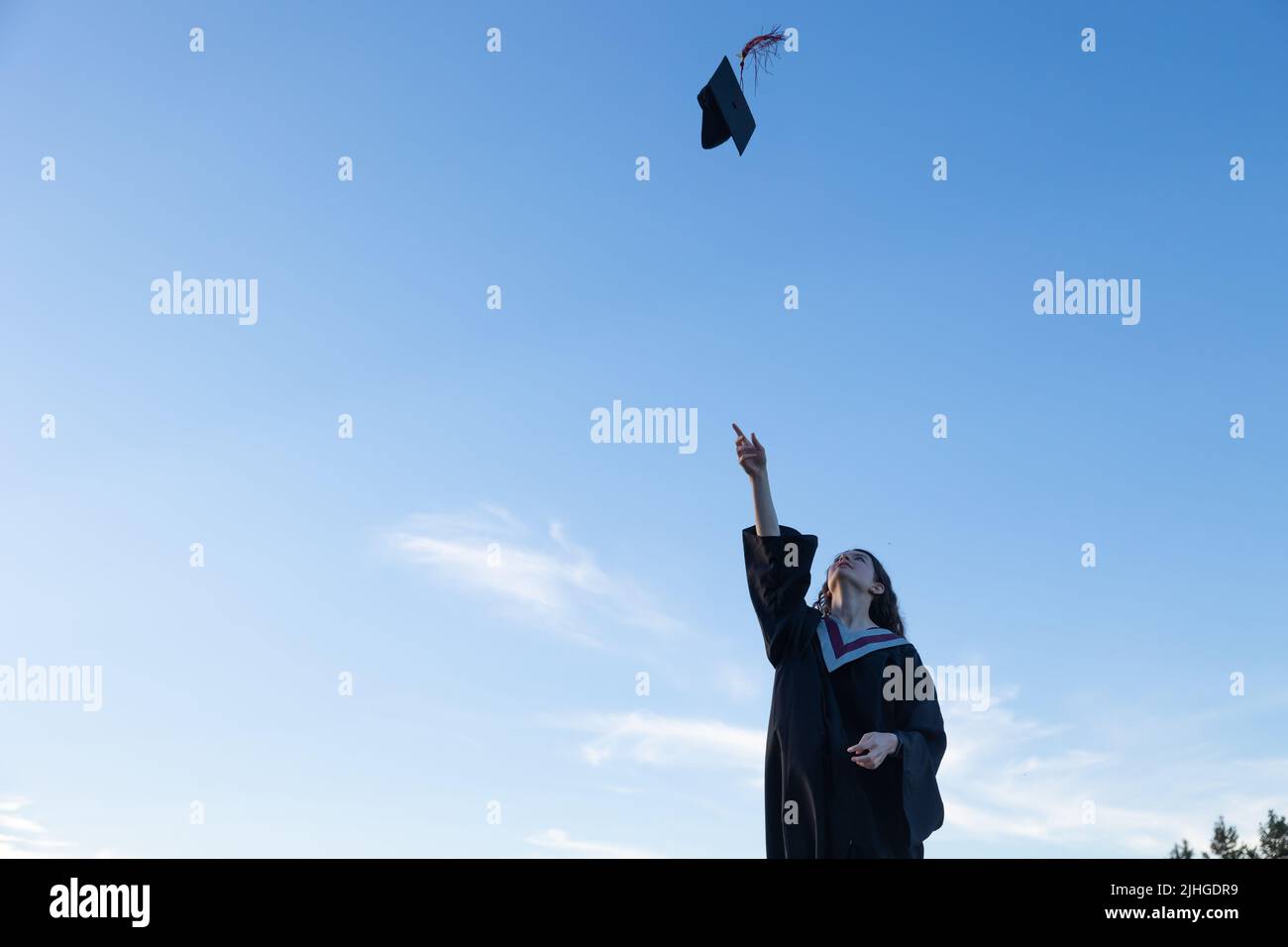 Junge Frau in Abschlusskleid, die ihre Mütze und ihr Mörtelbrett in die Luft wirft, mit einem blauen Himmel im Hintergrund Stockfoto