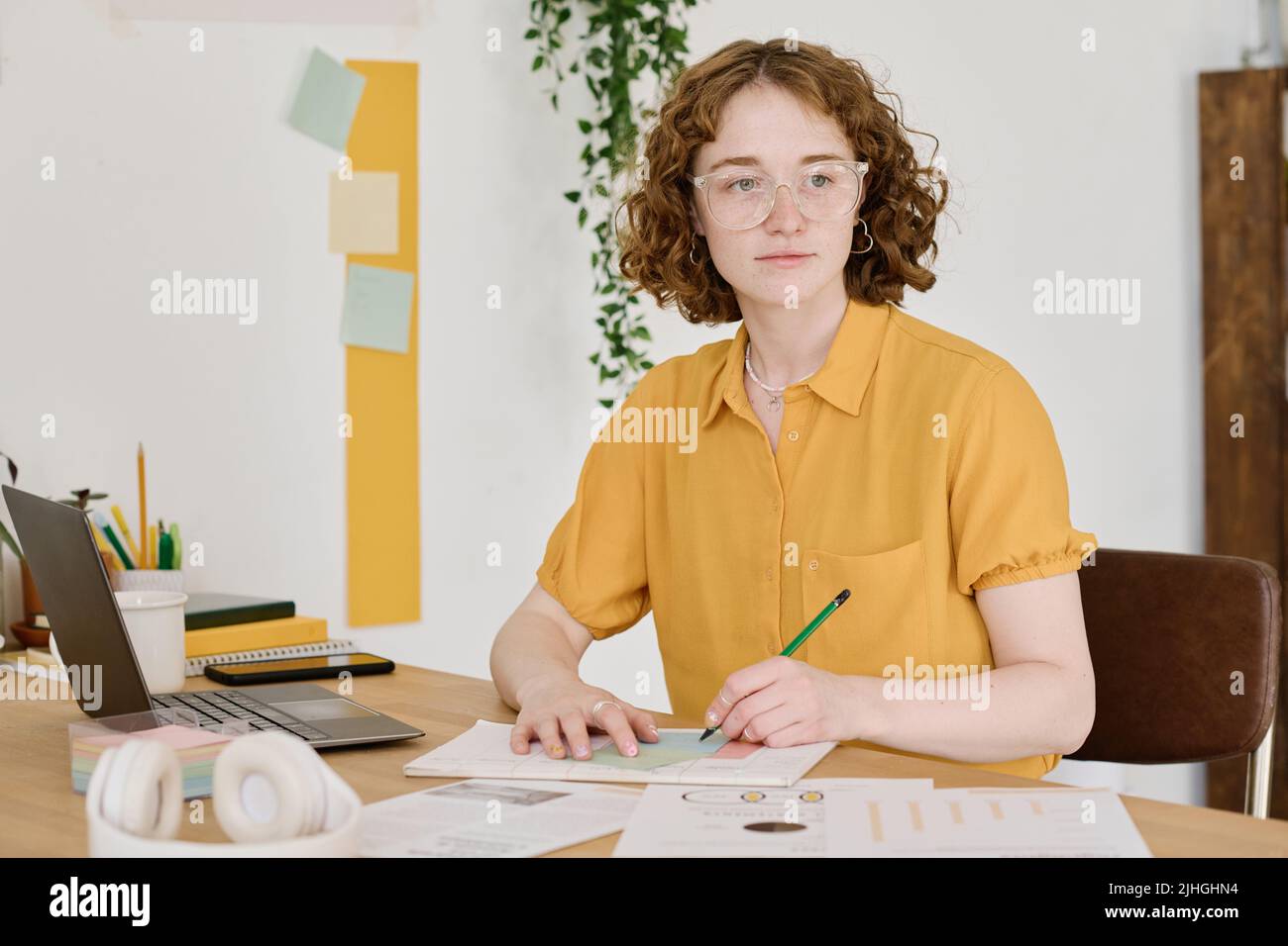 Junge kreative Frau in Brillen und gelben Blusen Zeichentwürfe auf Papier, während sie am Arbeitsplatz sitzt und nach vorne blickt Stockfoto