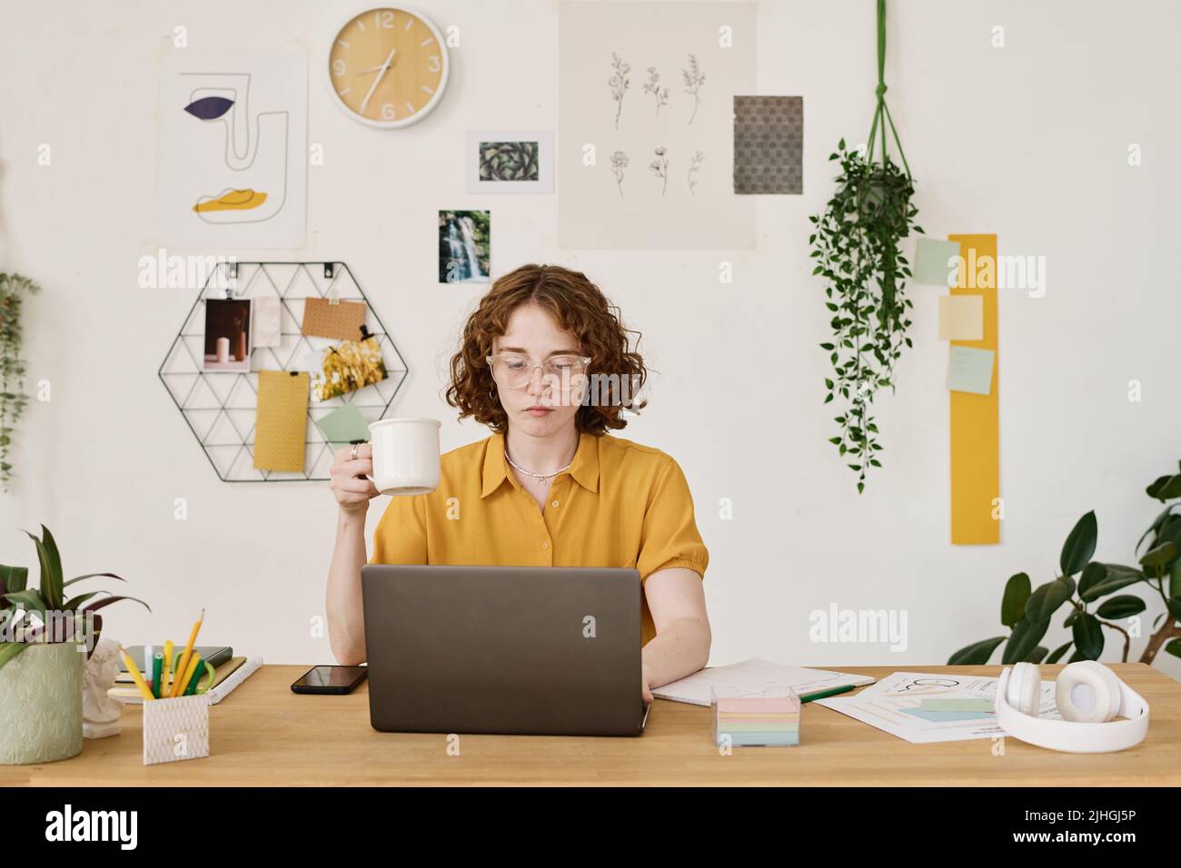 Junge, seriöse Freiberuflerin mit einer Tasse Tee, die am Arbeitsplatz vor dem Laptop sitzt und nach Online-Informationen für das Projekt sucht Stockfoto