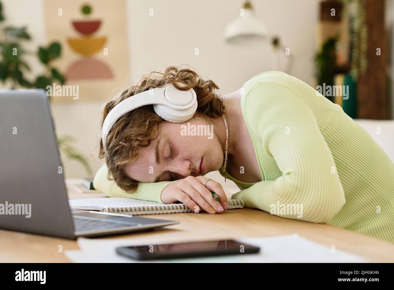 Junge, müde Studentin mit Kopfhörern und Casualwear, die den Kopf auf dem Copybook hält, während sie vor dem Laptop am Schreibtisch lappt Stockfoto