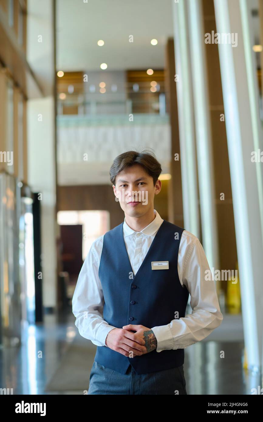 Der junge Hotelmanager oder Hotelpagen in Uniform steht vor der Kamera auf dem Flur oder in der Lounge und wartet auf neue Gäste Stockfoto