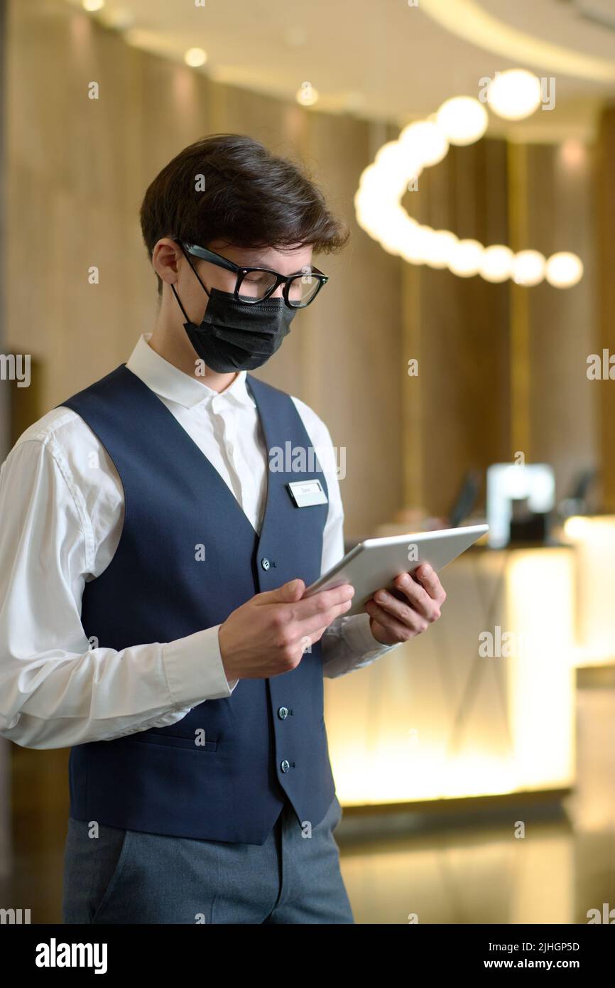 Männlicher Rezeptionist in Uniform und Schutzmaske, der Online-Reservierungen von Hotelzimmern überprüft, während er in der Lounge steht Stockfoto