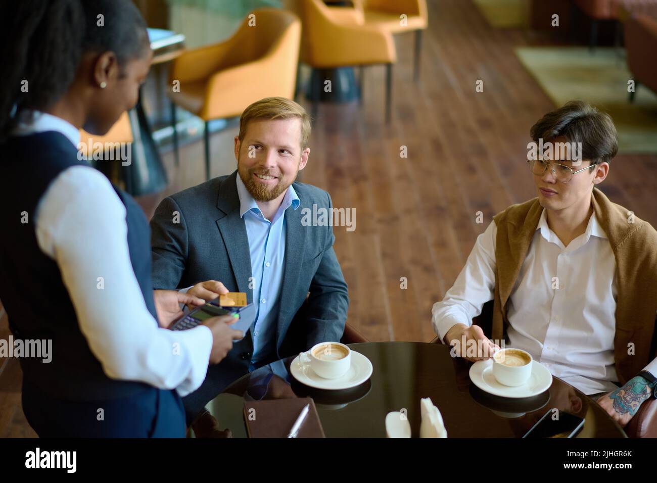 Glücklicher junger Geschäftsmann, der mit Kreditkarte für Kaffee in der Lounge oder im Café des Hotels bezahlt hat, während er die Managerin mit dem Zahlungsterminal ansah Stockfoto