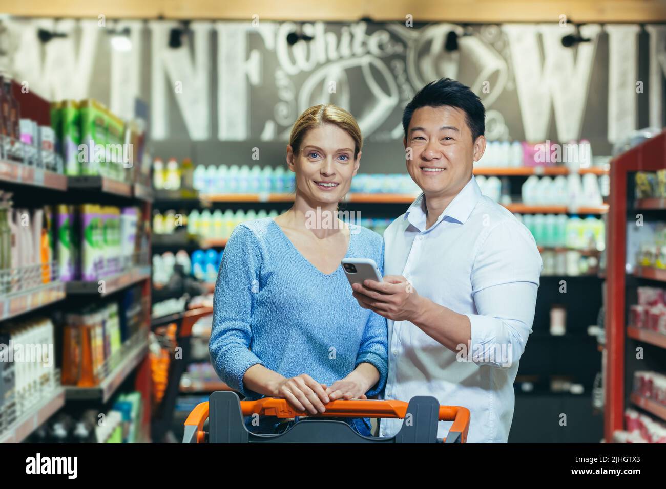 Familienportrait eines Paares beim Einkaufen im Supermarkt, lächelnd und mit Blick auf die Kamera, Käufer halten das Telefon in den Händen und nutzen die Rabatt-App Stockfoto