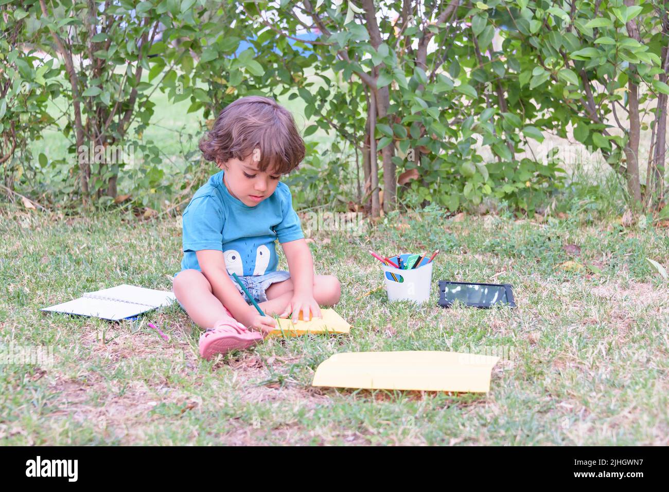 Der kleine Junge verwendet einen Zauberstift, um Bilder in einem Buch auf einem Holztisch im Park mit Gesichtsausdruck der Entschlossenheit zu zeichnen. Stockfoto