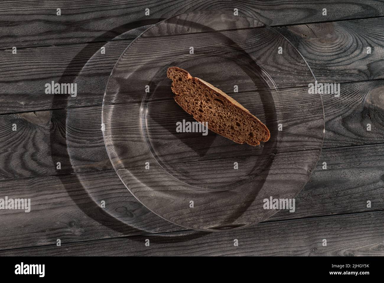 Braunes kleines Stück Brot auf großer Glasplatte auf schwarzem Holztisch. Dunkles HDR-Foto. Einfache Lebensmittel, magere Lebensmittel, Armut und Unterernährung Stockfoto
