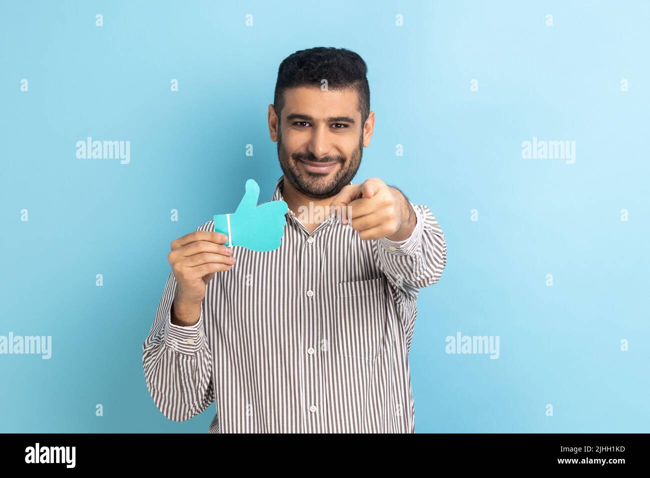 Bärtiger Geschäftsmann hält und zeigt ein Schild in Papierform, zeigt mit einem dutzenden Lächeln auf die Kamera und trägt ein gestreiftes Hemd. Innenaufnahme des Studios isoliert auf blauem Hintergrund. Stockfoto