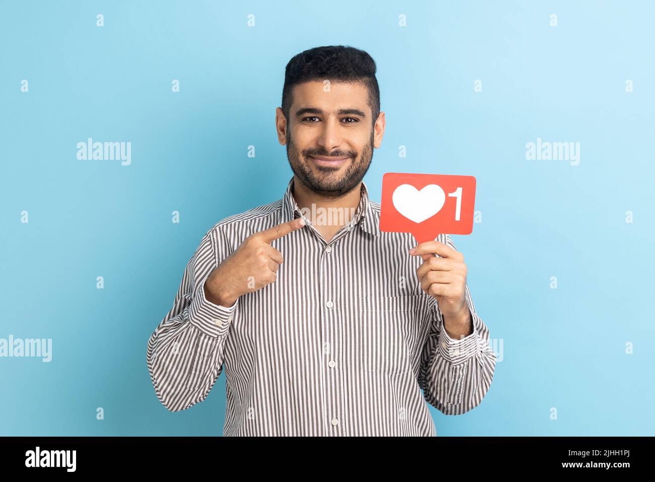 Internet-Blogging. Porträt eines fröhlichen Mannes, der wie ein Symbol auf das Herz zeigt und empfiehlt, auf die Social-Media-Schaltfläche zu klicken und ein gestreiftes Hemd zu tragen. Innenaufnahme des Studios isoliert auf blauem Hintergrund. Stockfoto