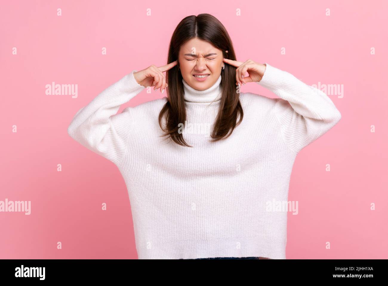 Porträt einer unglücklichen schönen Frau stehend und schloss ihre Ohren mit der Hand und wollen nicht hören, trägt weißen lässigen Stil Pullover. Innenaufnahme des Studios isoliert auf rosa Hintergrund. Stockfoto