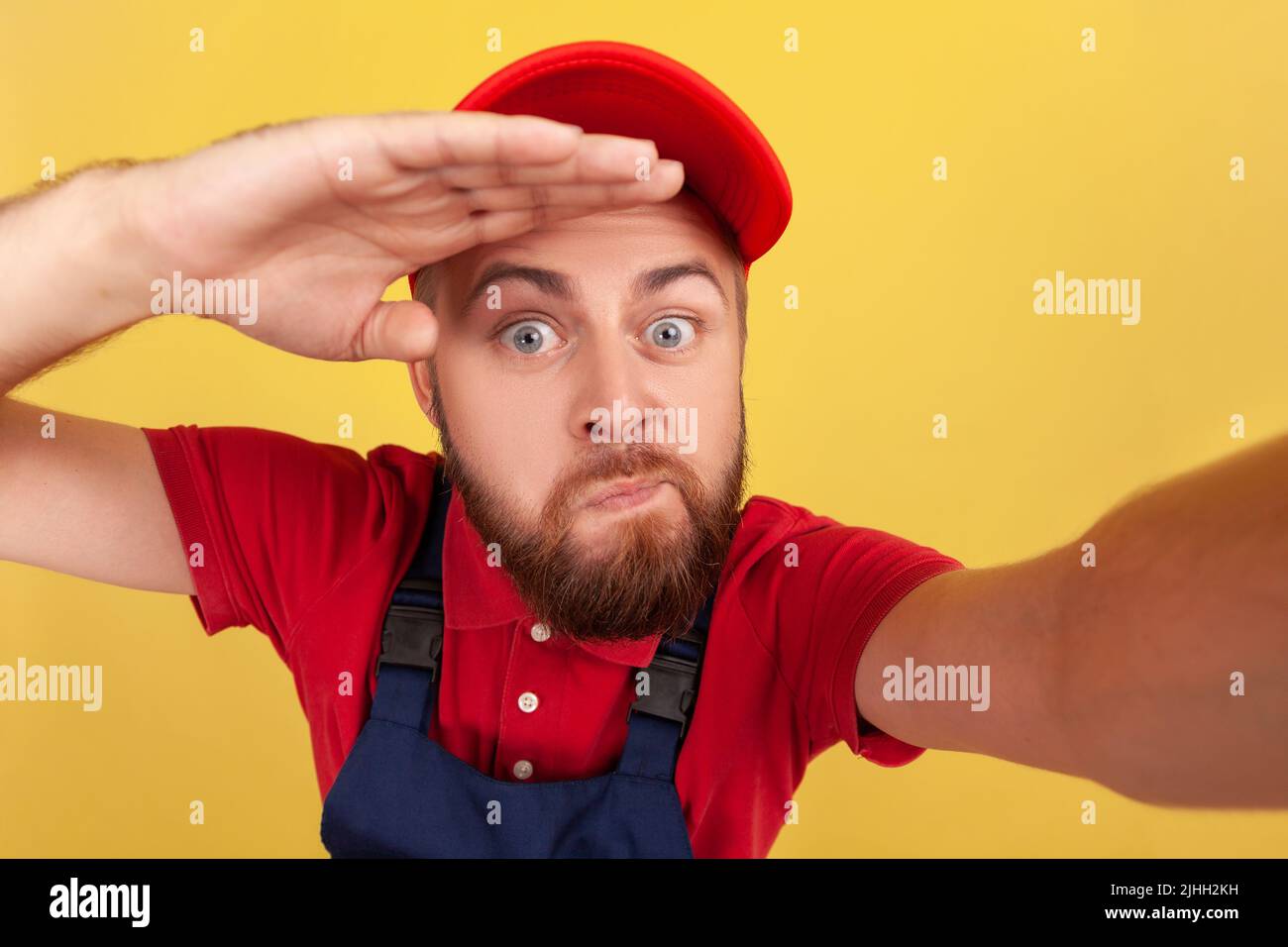 Lustiger Arbeiter mit blauen Overalls, der Selfie macht, mit der Hand über dem Kopf weit schaut, mit lächerlicher Ausdruckskraft POV, Standpunkt des Fotos. Innenaufnahme des Studios isoliert auf gelbem Hintergrund. Stockfoto