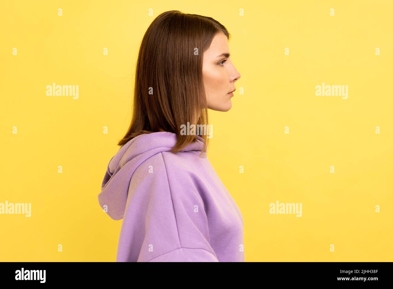 Seitenansicht einer jungen, seriös unlächelnden, strengen Frau, die mit einem ruhigen, aufmerksamen Gesichtsausdruck die Kamera anschaut und einen violetten Hoodie trägt. Innenaufnahme des Studios isoliert auf gelbem Hintergrund. Stockfoto