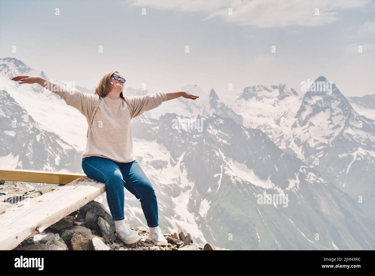 Glücklich attraktive Frau in Sonnenbrille sitzt auf einem Felsen mit erhobenen Händen und Blick in den Himmel. Fröhlicher Tourist in den Bergen. Schneeberggipfel auf dem Rücken Stockfoto