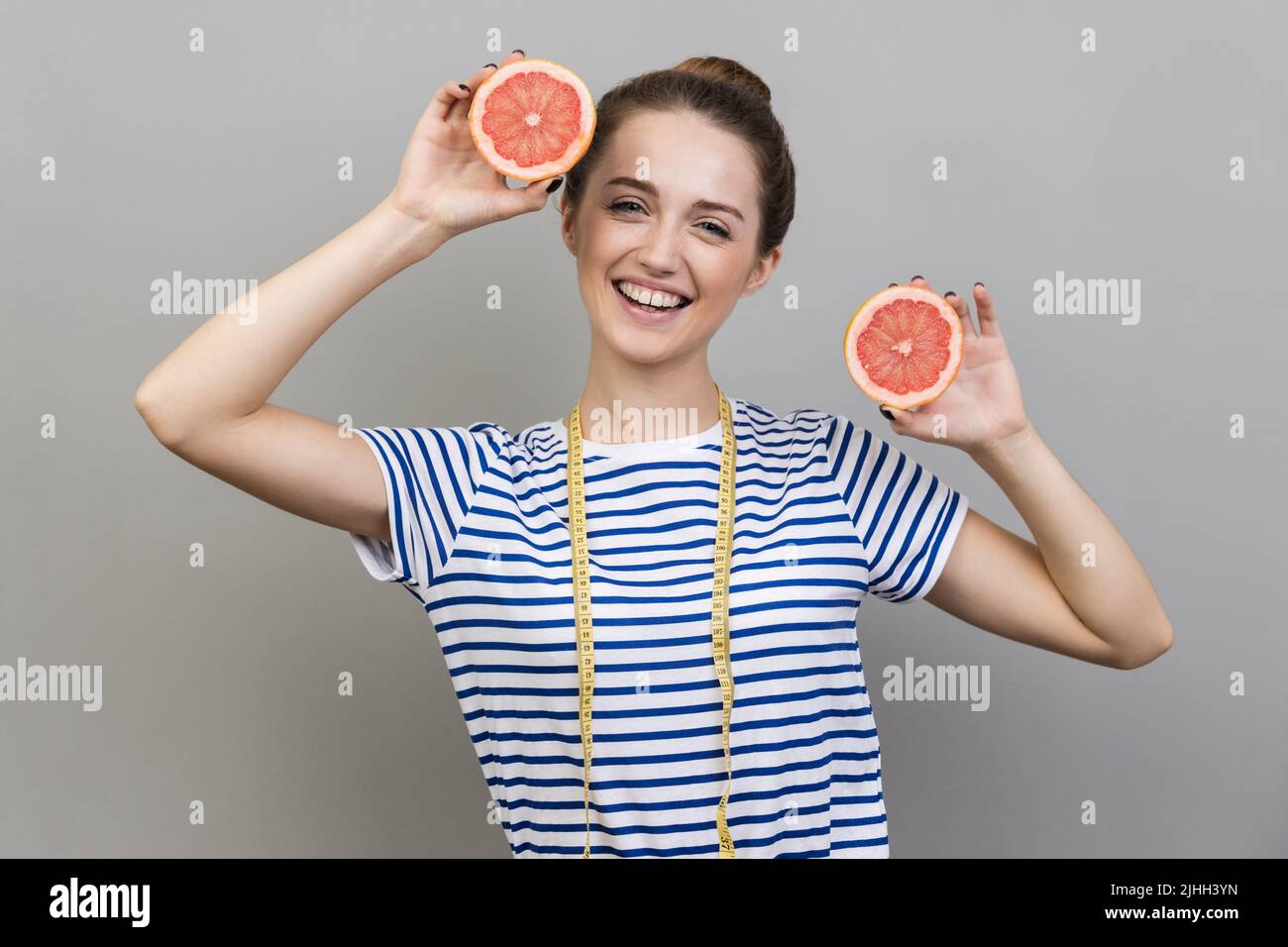 Porträt einer fröhlichen, optimistischen Frau, die gestreiftes T-Shirt trägt und mit Grapefruitscheiben und Maßband steht, lächelnd auf die Kamera. Innenaufnahme auf grauem Hintergrund. Stockfoto